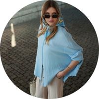 A woman wears a light blue, oversized muslin blouse with wide sleeves, paired with beige pants and a patterned scarf – blue muslin blouses by SASSYCLASSY